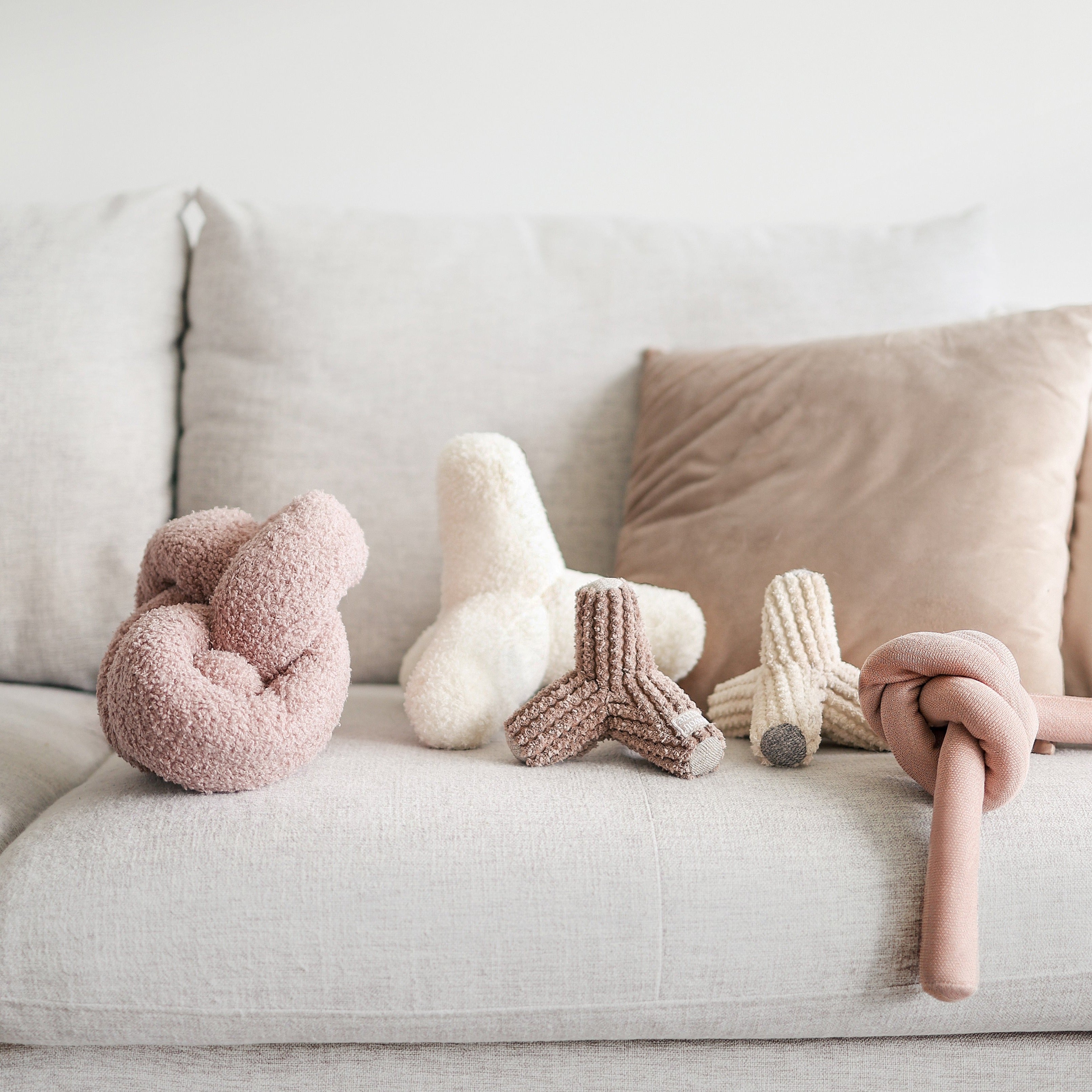 Five plush dog toys rest on a white couch, showcasing various textures and knot shapes for playful chewing.