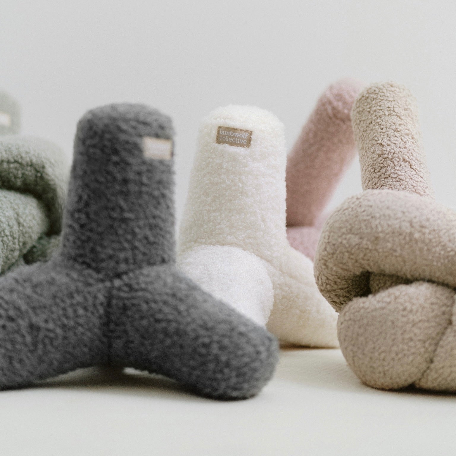 Four soft, knot-shaped pillows in charcoal, cream, dusty rose, and beige rest on a white surface. 
