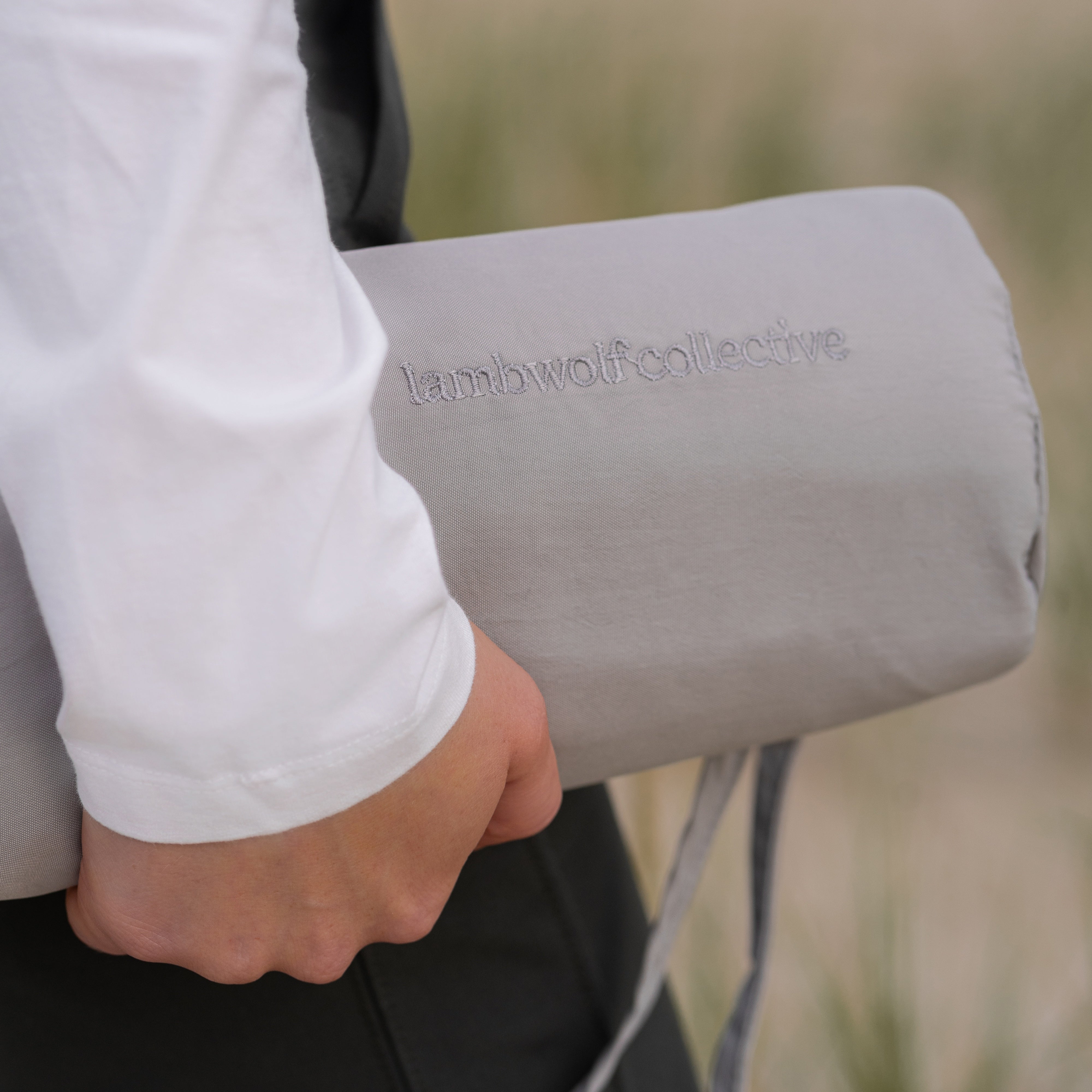 Woman carries a rolled-up gray yoga mat with the embroidered brand name "Lambwolf Collective". 
