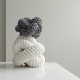 A gray and white chunky knit sculpture sits on a white table, adding a touch of texture to a minimalist room. 
