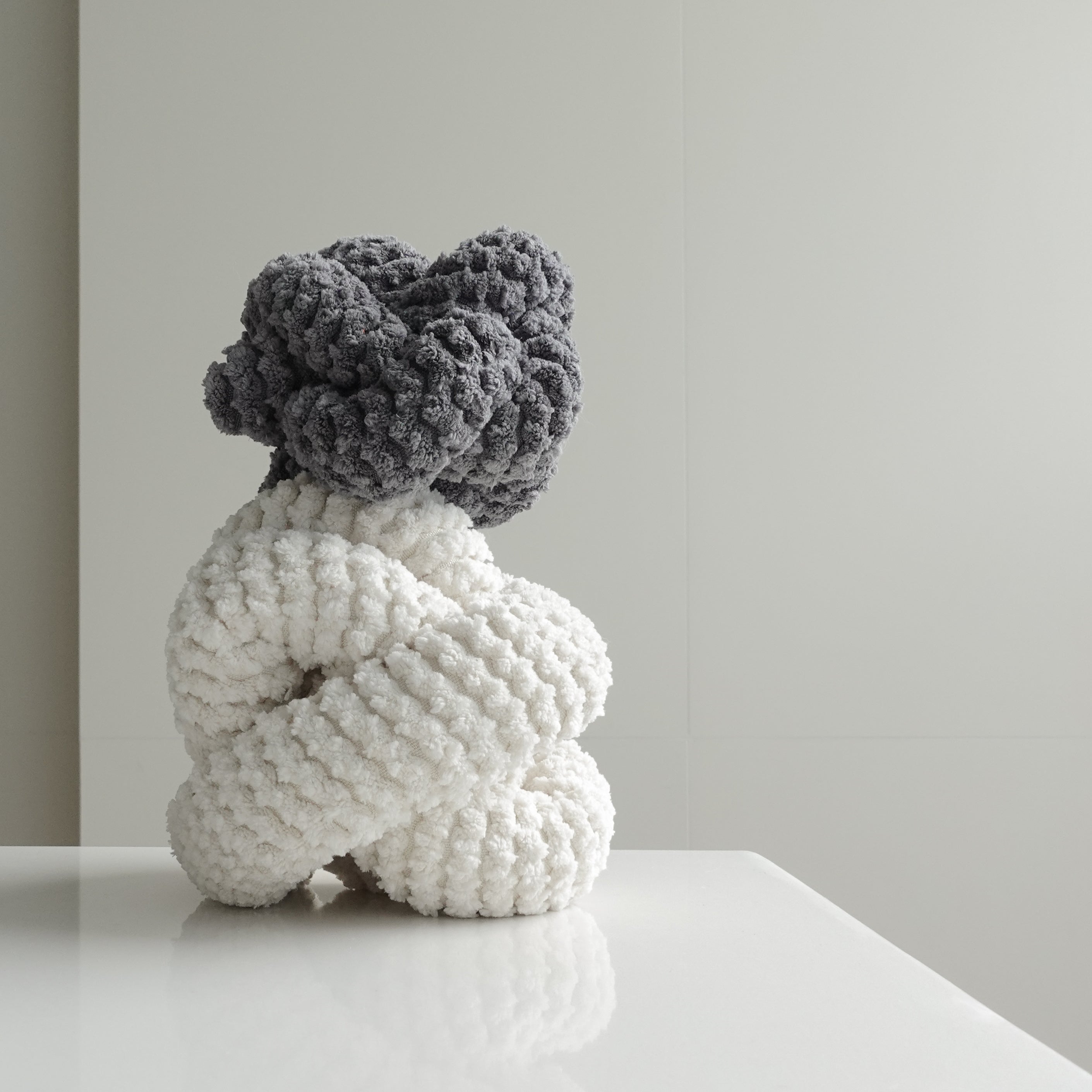 A gray and white chunky knit sculpture sits on a white table, adding a touch of texture to a minimalist room. 
