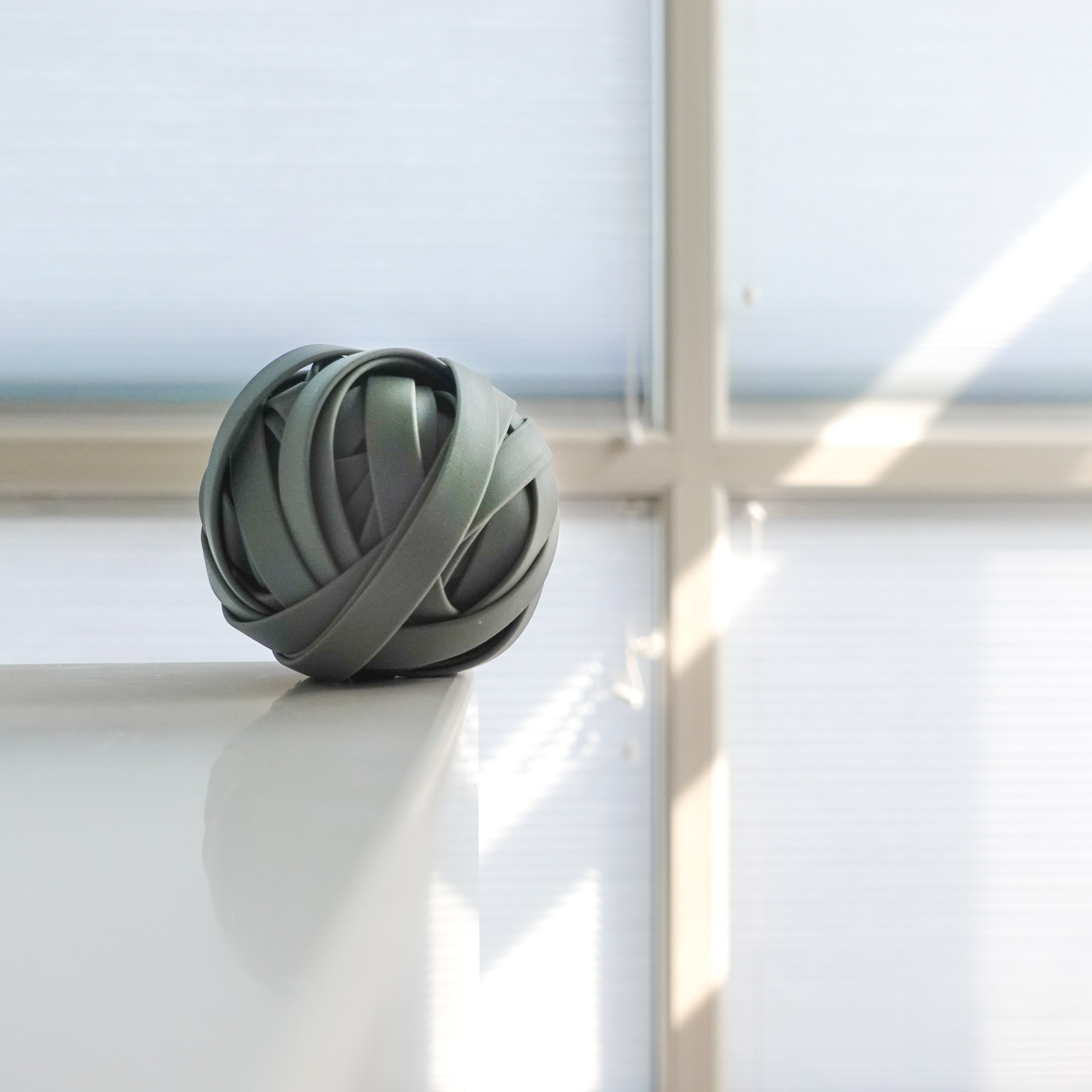 A green rubber band ball sits on a white table, illuminated by soft window light. 
