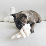 A brindle French Bulldog chews on a soft, knotted plush toy while relaxing on a light grey couch. 
