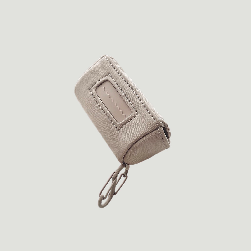 A beige dog poop bag holder hangs from a carabiner clip. The dispenser has a clear window on the front to see remaining bags. 
