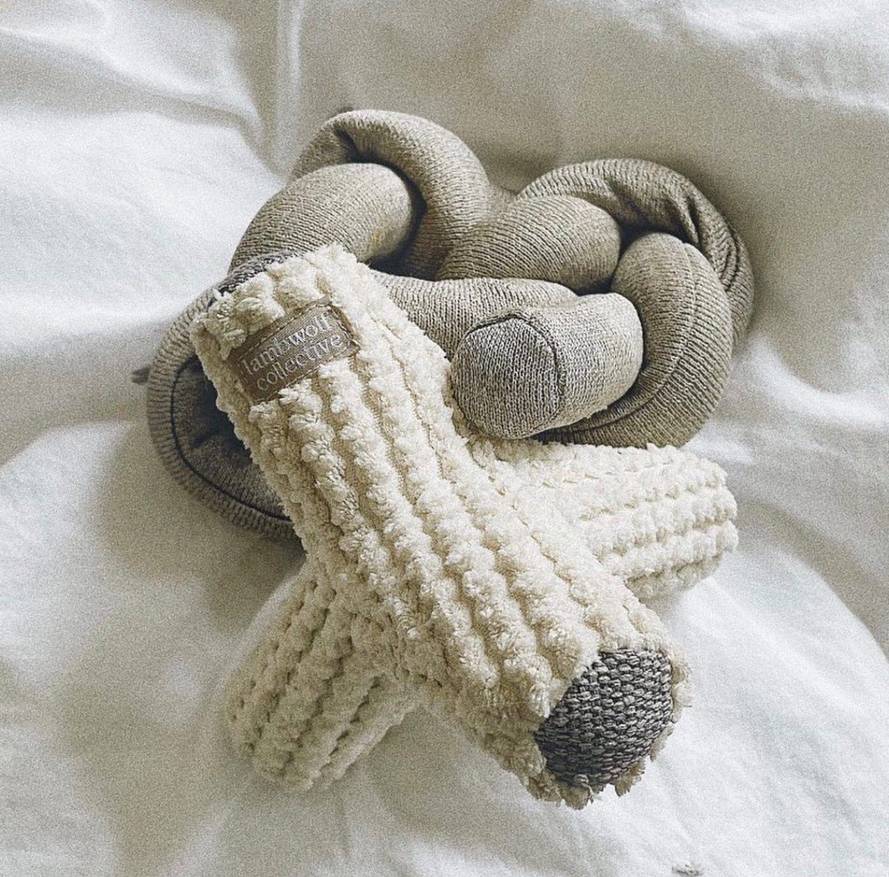 Plush knotted baby lounger in oatmeal color rests on white sheets. A soft, textured white baby toy with a nubby texture lays on top. 
