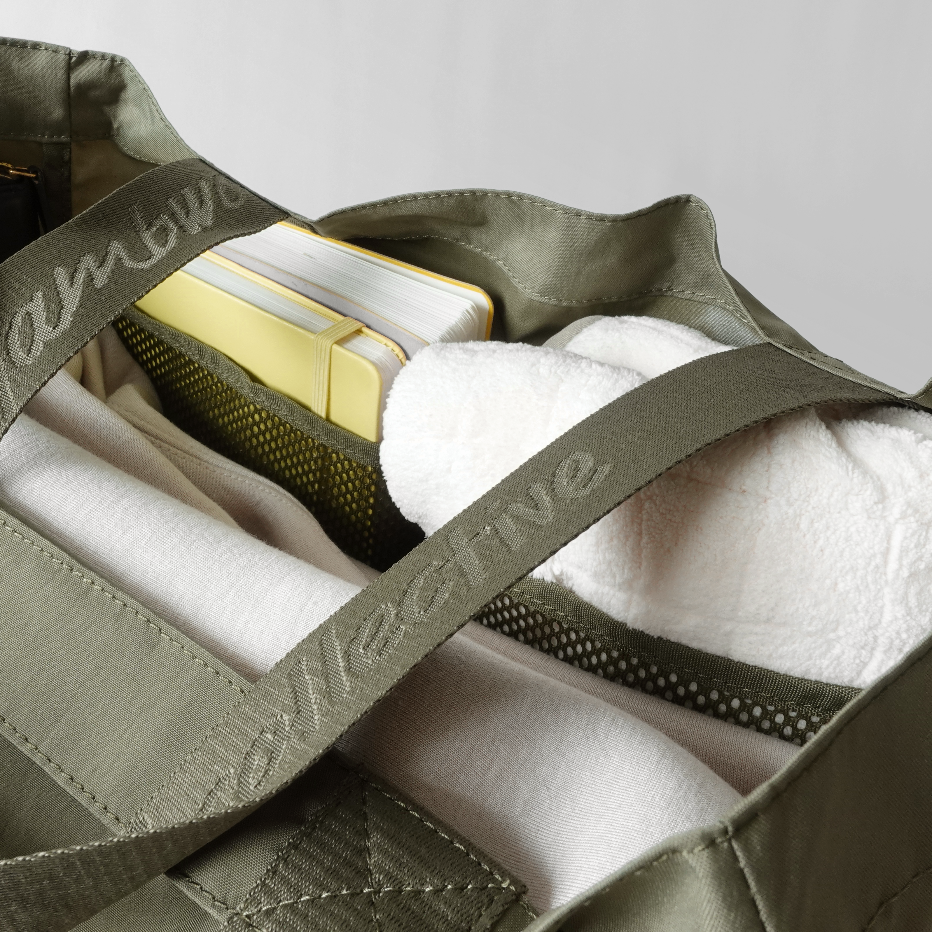 Open olive green tote bag holds a yellow notebook, white clothing items, and a white sherpa jacket. 
