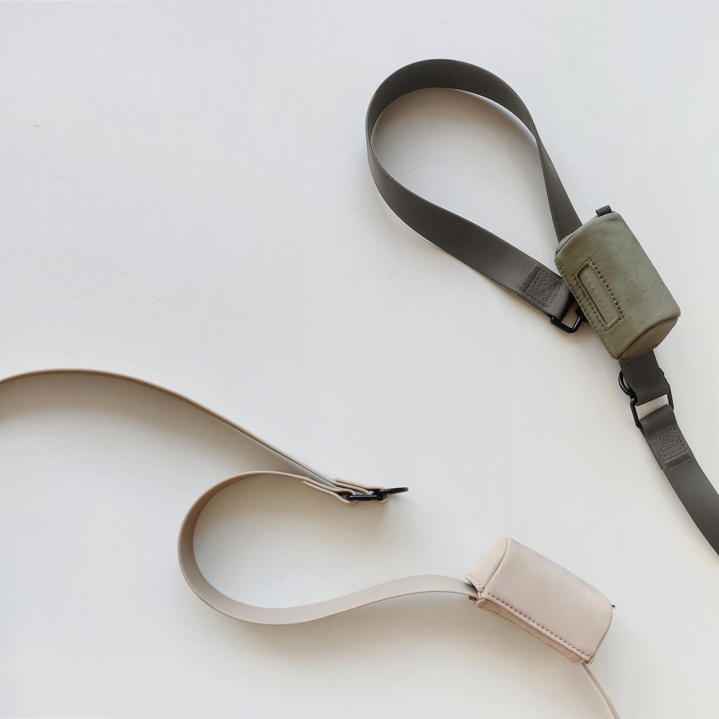 Two minimalist crossbody bags in beige and olive green lie on a white surface. 
