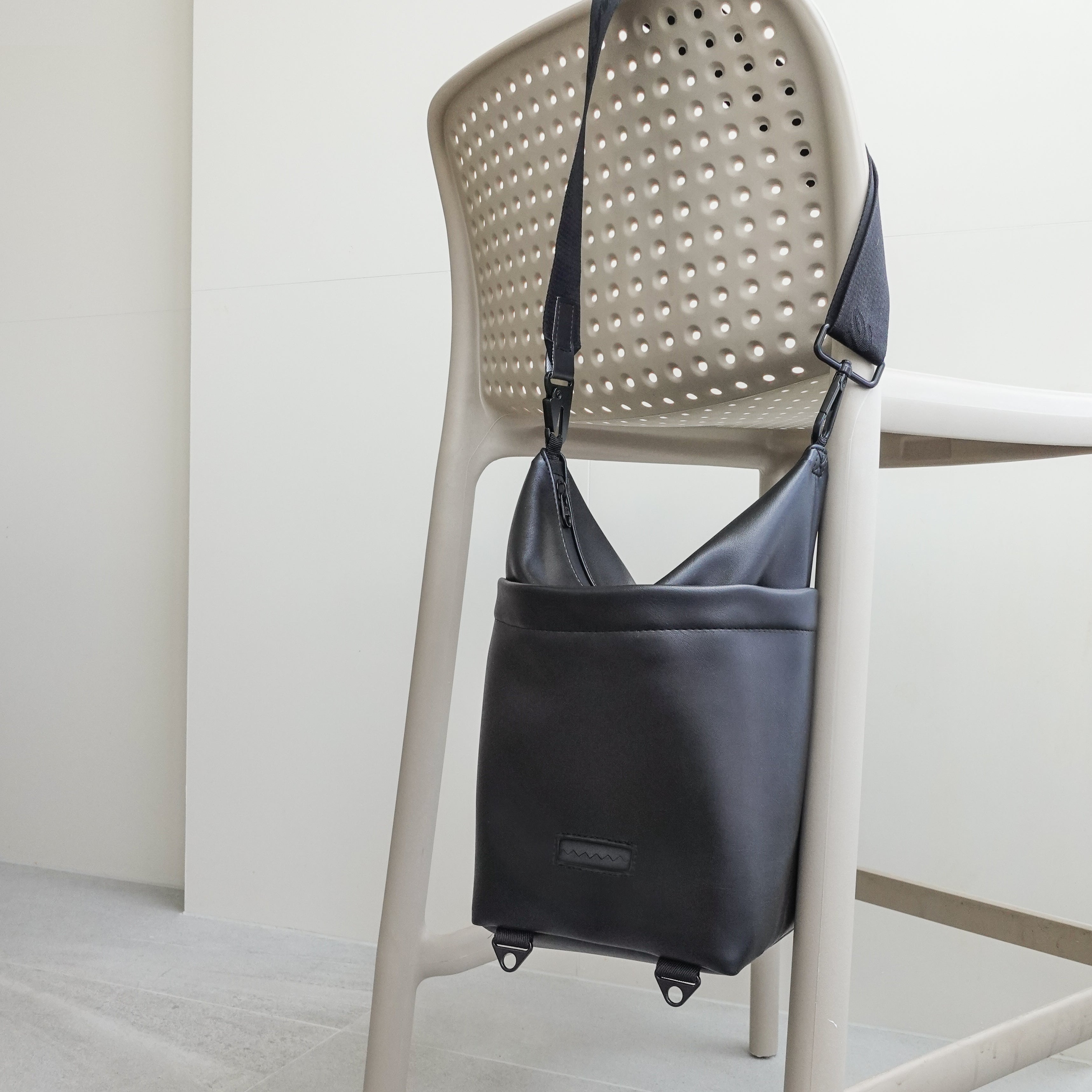 Black leather crossbody bag hangs from a modern chair, showcasing its sleek design and adjustable strap. 
