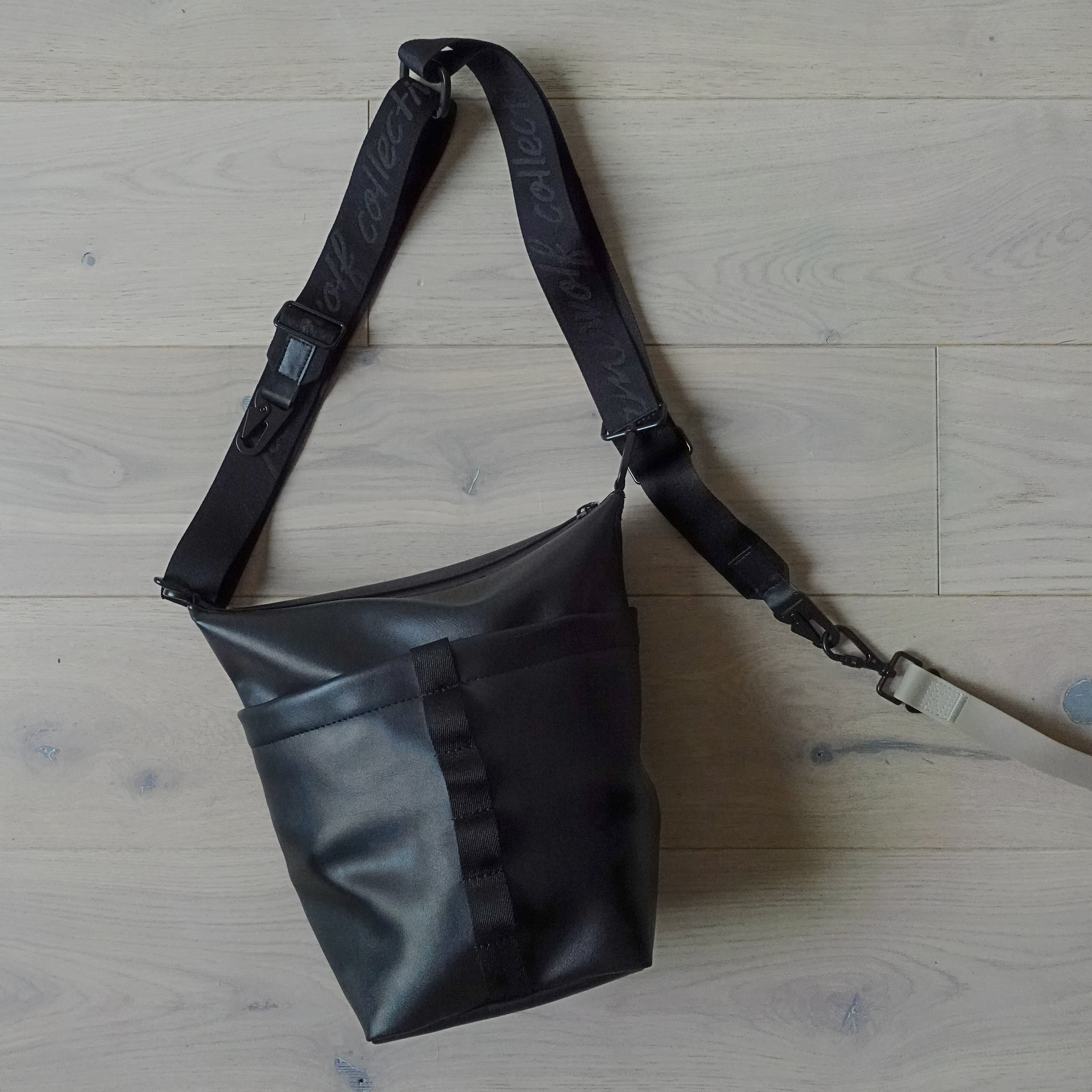 Black crossbody bag with adjustable strap lays on light wood floor. 
