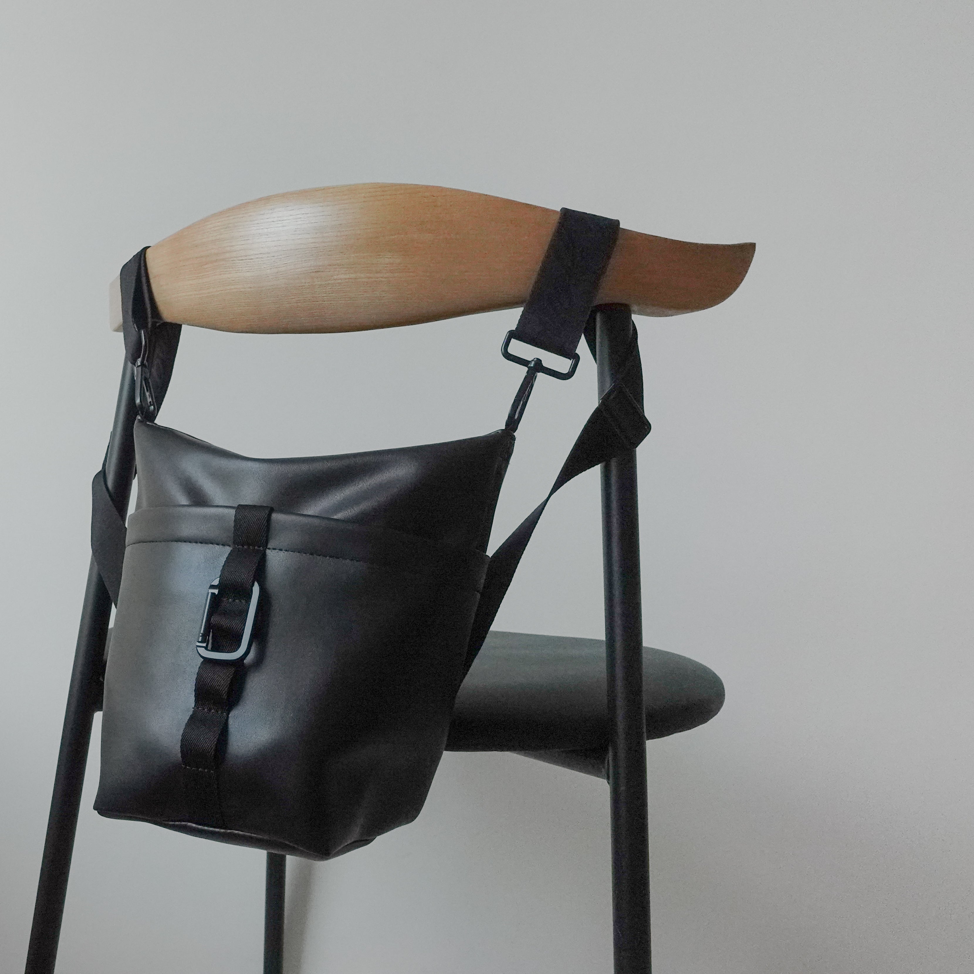 Black crossbody bag with adjustable strap hangs on the back of a modern chair. 
